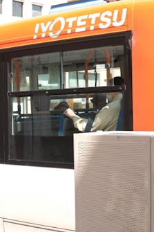An orange and white bus with the name 'IYOTETSU' on the side. Through the window, passengers are visible sitting inside. The surroundings outside the bus include part of a building and a metal structure.