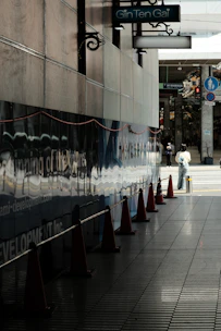 A row of orange traffic cones line a dark tiled walkway next to a wall with reflective panels displaying text and a construction-related advertisement. Above, a sign reads 'Gin Ten Gai' in blue on a dark signboard. In the background, a person stands on the sidewalk, partially visible, suggesting an urban environment.