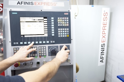A person operates a control panel with various buttons, switches, and a digital screen displaying data. The background features a machine labeled AFINIS EXPRESS.