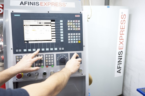 A person operates a control panel with various buttons, switches, and a digital screen displaying data. The background features a machine labeled AFINIS EXPRESS.