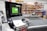 A computerized workstation in a workshop, featuring a monitor displaying green digital patterns and a keyboard and mouse in the foreground. Behind the workstation, there are shelves filled with various tools and materials, including rolls of fabric or paper.