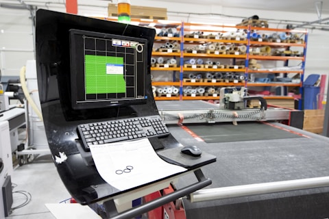 A computerized workstation in a workshop, featuring a monitor displaying green digital patterns and a keyboard and mouse in the foreground. Behind the workstation, there are shelves filled with various tools and materials, including rolls of fabric or paper.