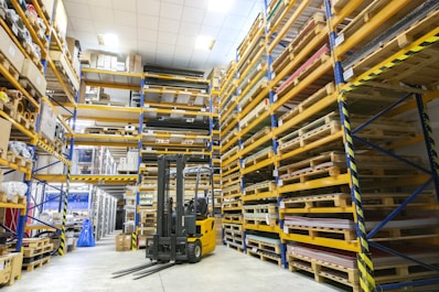 A spacious warehouse with tall shelves filled with various boxes and pallets. A yellow forklift is parked in the aisle, surrounded by organized inventory. The environment appears clean and well-lit, with items neatly stacked on metal racks.