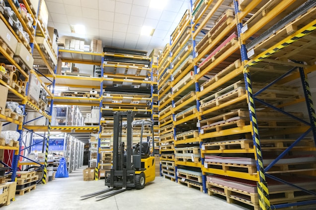 A spacious warehouse with tall shelves filled with various boxes and pallets. A yellow forklift is parked in the aisle, surrounded by organized inventory. The environment appears clean and well-lit, with items neatly stacked on metal racks.