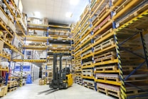 A spacious warehouse with tall shelves filled with various boxes and pallets. A yellow forklift is parked in the aisle, surrounded by organized inventory. The environment appears clean and well-lit, with items neatly stacked on metal racks.