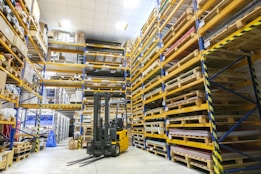 A spacious warehouse with tall shelves filled with various boxes and pallets. A yellow forklift is parked in the aisle, surrounded by organized inventory. The environment appears clean and well-lit, with items neatly stacked on metal racks.