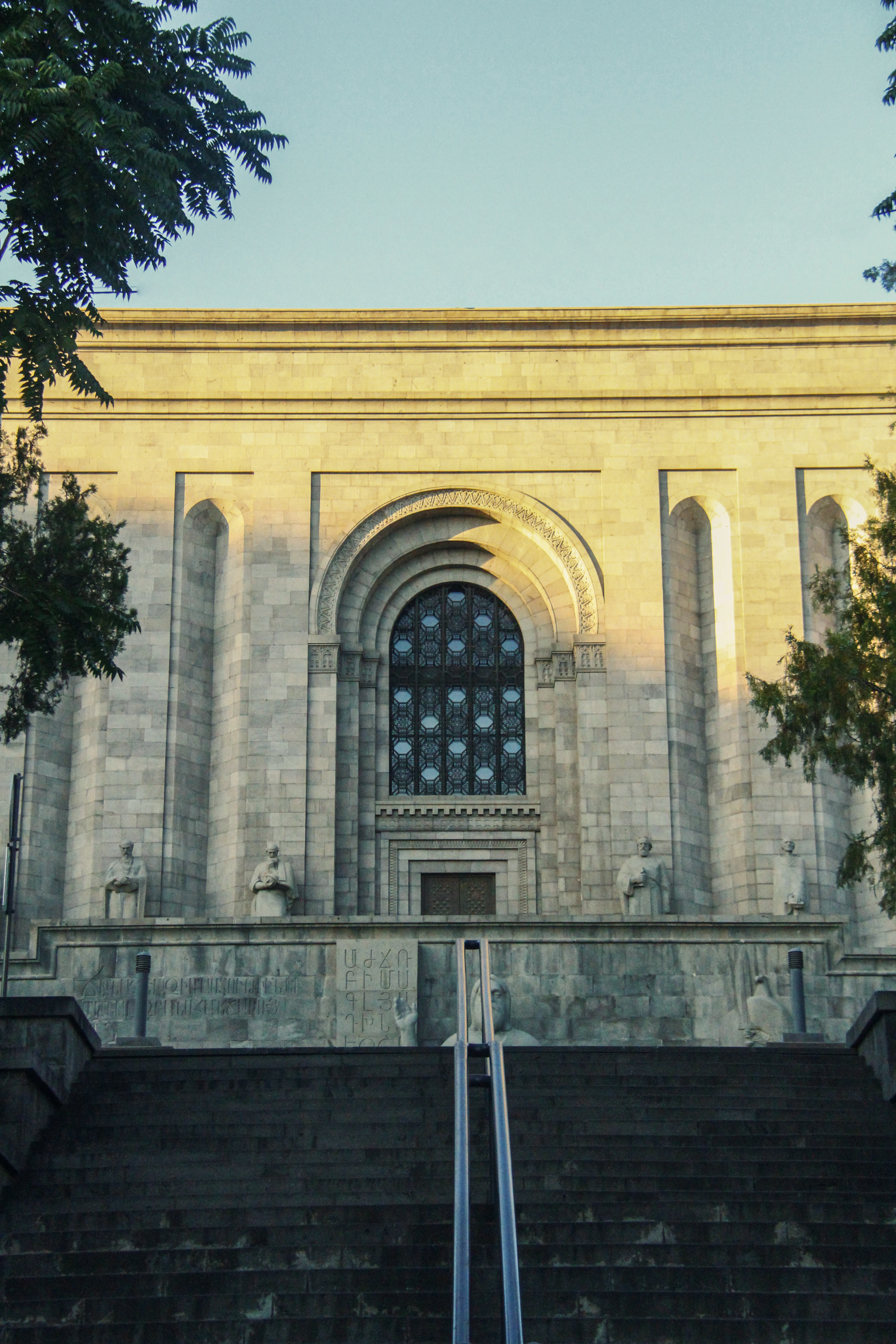 The Matenadaran in Yerevan at the top of its long flight of steps, a monumental grey basalt facade with a tall arched stained-glass window, statues of Armenian scholars seated in niches and carved Armenian inscription across the wall, the upper stonework catching late sun