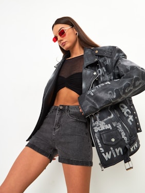 A person poses confidently wearing a black leather jacket with text designs, paired with a sheer black top and high-waisted gray denim shorts. Red-tinted sunglasses add a bold fashion statement.