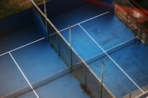 An overhead view of two adjacent blue tennis or paddle courts with a slightly worn surface. The courts are separated by fencing and have white lines marking the play areas. Surrounded by grass and tiles, the setting suggests an outdoor sports complex.