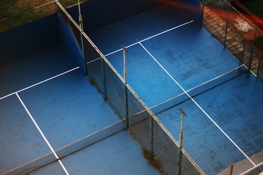 An overhead view of two adjacent blue tennis or paddle courts with a slightly worn surface. The courts are separated by fencing and have white lines marking the play areas. Surrounded by grass and tiles, the setting suggests an outdoor sports complex.