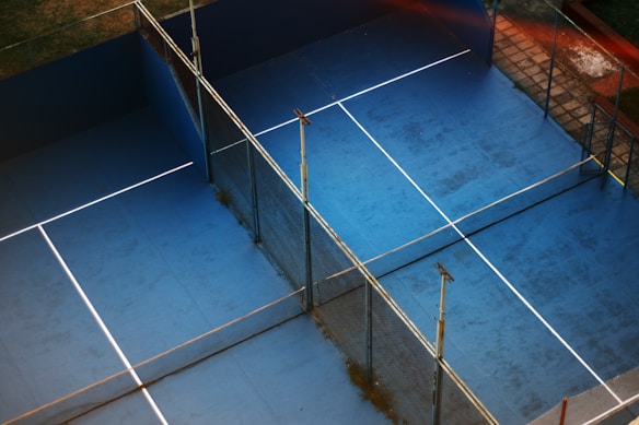 An overhead view of two adjacent blue tennis or paddle courts with a slightly worn surface. The courts are separated by fencing and have white lines marking the play areas. Surrounded by grass and tiles, the setting suggests an outdoor sports complex.