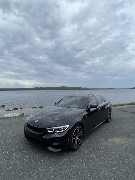 A sleek black luxury car shining under the sun near a coastal shoreline with gentle waves.
