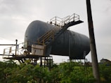 A large, industrial cylindrical tank is elevated on metal supports, with a staircase and railing attached to its side. The structure appears weathered and is set against a cloudy sky. Vegetation is visible in the foreground, with an electrical pole nearby.