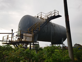 A large, industrial cylindrical tank is elevated on metal supports, with a staircase and railing attached to its side. The structure appears weathered and is set against a cloudy sky. Vegetation is visible in the foreground, with an electrical pole nearby.
