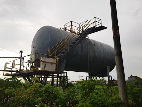 A large, industrial cylindrical tank is elevated on metal supports, with a staircase and railing attached to its side. The structure appears weathered and is set against a cloudy sky. Vegetation is visible in the foreground, with an electrical pole nearby.