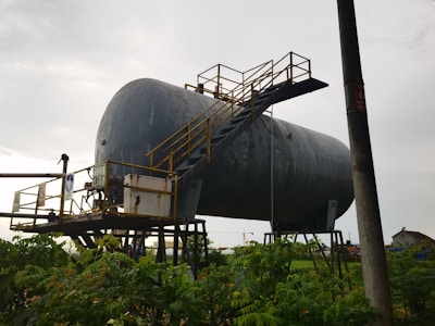 A large, industrial cylindrical tank is elevated on metal supports, with a staircase and railing attached to its side. The structure appears weathered and is set against a cloudy sky. Vegetation is visible in the foreground, with an electrical pole nearby.