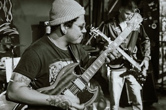 A musician wearing a beanie and glasses plays an electric guitar, concentrating intensely on the music. Tattoos are visible on the musician's arms. In the background, another person plays a bass guitar, and a drum set can be partially seen.
