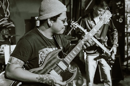 A musician wearing a beanie and glasses plays an electric guitar, concentrating intensely on the music. Tattoos are visible on the musician's arms. In the background, another person plays a bass guitar, and a drum set can be partially seen.