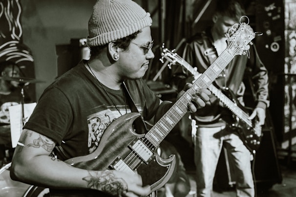 A musician wearing a beanie and glasses plays an electric guitar, concentrating intensely on the music. Tattoos are visible on the musician's arms. In the background, another person plays a bass guitar, and a drum set can be partially seen.