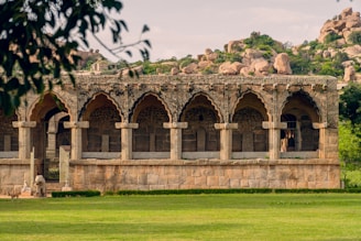 A historic stone structure with arched doorways and intricate stonework, set in a natural landscape. The building is surrounded by greenery and rocky hills are visible in the background. A large lawn stretches in front of the structure, and a statue can be seen nearby.