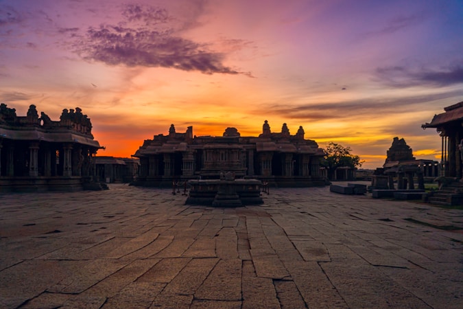A historic temple complex under a vibrant sunset sky with silhouettes of ancient stone structures. The courtyard showcases stone pavements leading towards intricately carved temples.