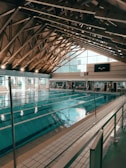 An indoor swimming pool with several lanes is housed under a high wooden ceiling with exposed trusses. Large windows allow natural light to fill the space. Poolside seating and time displays are visible around the pool area.
