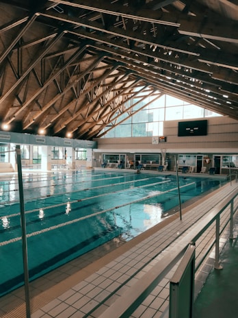 An indoor swimming pool with several lanes is housed under a high wooden ceiling with exposed trusses. Large windows allow natural light to fill the space. Poolside seating and time displays are visible around the pool area.
