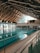 An indoor swimming pool with several lanes is housed under a high wooden ceiling with exposed trusses. Large windows allow natural light to fill the space. Poolside seating and time displays are visible around the pool area.