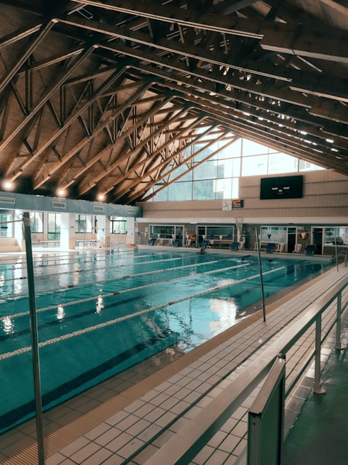 An indoor swimming pool with several lanes is housed under a high wooden ceiling with exposed trusses. Large windows allow natural light to fill the space. Poolside seating and time displays are visible around the pool area.