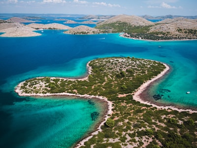 An aerial view of a large, heart-shaped island surrounded by clear turquoise waters. The island is covered with lush greenery and patches of rocky terrain. The surrounding water varies in shades of blue, indicating different depths. In the background, several smaller islands and hills can be seen against a blue sky with scattered clouds.