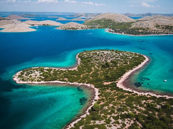 An aerial view of a large, heart-shaped island surrounded by clear turquoise waters. The island is covered with lush greenery and patches of rocky terrain. The surrounding water varies in shades of blue, indicating different depths. In the background, several smaller islands and hills can be seen against a blue sky with scattered clouds.