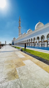 A grand mosque with a series of white domes and a tall minaret under a clear blue sky. The architecture features ornate columns with golden accents. The foreground consists of a marble pathway accompanied by a strip of green grass alongside a reflective body of water. A person is walking along the pathway.