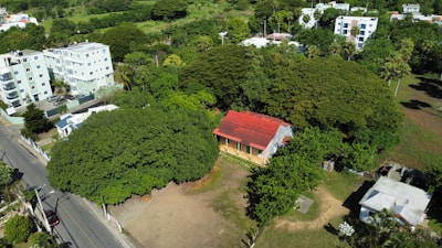 Aerial view of a residential area featuring several multi-story buildings surrounded by dense greenery. In the foreground, there is a small house with a red roof nestled among large trees.