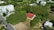 Aerial view of a residential area featuring several multi-story buildings surrounded by dense greenery. In the foreground, there is a small house with a red roof nestled among large trees.