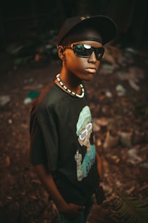 A young person wearing a black cap, sunglasses, and a black shirt with a graphic print. A necklace with white beads complements the look. The background is blurred and appears to be outdoors with some leaves and debris on the ground.