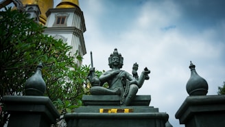 A statue of a multi-armed deity is prominently displayed in the center, surrounded by ornate structures and greenery. The statue is seated on a pedestal with intricate carvings, holding various objects in its hands. In the background, there is a temple-like structure with golden and white detailing, set against a cloudy sky.