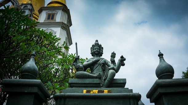 A statue of a multi-armed deity is prominently displayed in the center, surrounded by ornate structures and greenery. The statue is seated on a pedestal with intricate carvings, holding various objects in its hands. In the background, there is a temple-like structure with golden and white detailing, set against a cloudy sky.