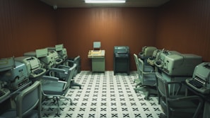 A vintage computer room containing multiple old-fashioned teletypes and office chairs arranged in a semi-circle around the walls. The room features wooden panel walls and a patterned black-and-white tiled floor, with fluorescent lighting overhead.