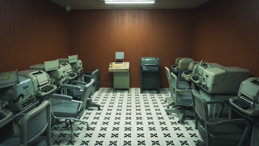 A vintage computer room containing multiple old-fashioned teletypes and office chairs arranged in a semi-circle around the walls. The room features wooden panel walls and a patterned black-and-white tiled floor, with fluorescent lighting overhead.