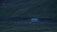 A bus travels along a winding road set against a backdrop of rolling, grassy hills during dusk or dawn. The landscape is shrouded in a dim, bluish hue, suggesting a serene and quiet atmosphere.