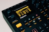 The image features a close-up view of a music production device, specifically an 8-voice digital drum computer and sampler. The display screen shows various settings and options, with numerous buttons, knobs, and controls visible around it. The color scheme is predominantly black with accents of orange and yellow. The brand name 'Digitakt' is prominently displayed below the screen.