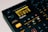 The image features a close-up view of a music production device, specifically an 8-voice digital drum computer and sampler. The display screen shows various settings and options, with numerous buttons, knobs, and controls visible around it. The color scheme is predominantly black with accents of orange and yellow. The brand name 'Digitakt' is prominently displayed below the screen.