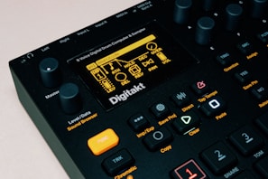 The image features a close-up view of a music production device, specifically an 8-voice digital drum computer and sampler. The display screen shows various settings and options, with numerous buttons, knobs, and controls visible around it. The color scheme is predominantly black with accents of orange and yellow. The brand name 'Digitakt' is prominently displayed below the screen.