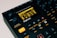 The image features a close-up view of a music production device, specifically an 8-voice digital drum computer and sampler. The display screen shows various settings and options, with numerous buttons, knobs, and controls visible around it. The color scheme is predominantly black with accents of orange and yellow. The brand name 'Digitakt' is prominently displayed below the screen.