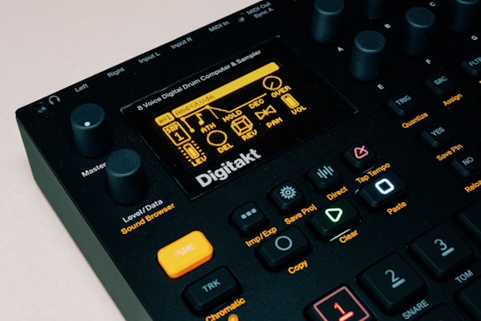 The image features a close-up view of a music production device, specifically an 8-voice digital drum computer and sampler. The display screen shows various settings and options, with numerous buttons, knobs, and controls visible around it. The color scheme is predominantly black with accents of orange and yellow. The brand name 