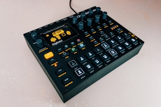 A black electronic music device with multiple buttons and knobs is set against a light background. The device has a digital display showing pixelated graphics. The buttons are illuminated in blue with black labels, and there are yellow accents on specific buttons. The device appears to be compact and designed for music production or manipulation.
