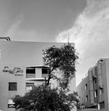 A black and white photograph of a building with a tree in the foreground. The building has a sign that reads 'restaurant' and features multiple windows and architectural details. The sky is slightly overcast, creating a muted atmosphere.