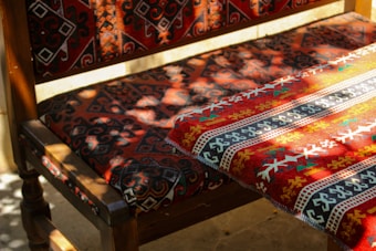 A wooden chair and table draped with richly patterned fabric featuring intricate geometric designs. The fabric predominantly displays a combination of red, black, white, and yellow colors, illuminated by sunlight filtering through leaves, creating a play of light and shadow.