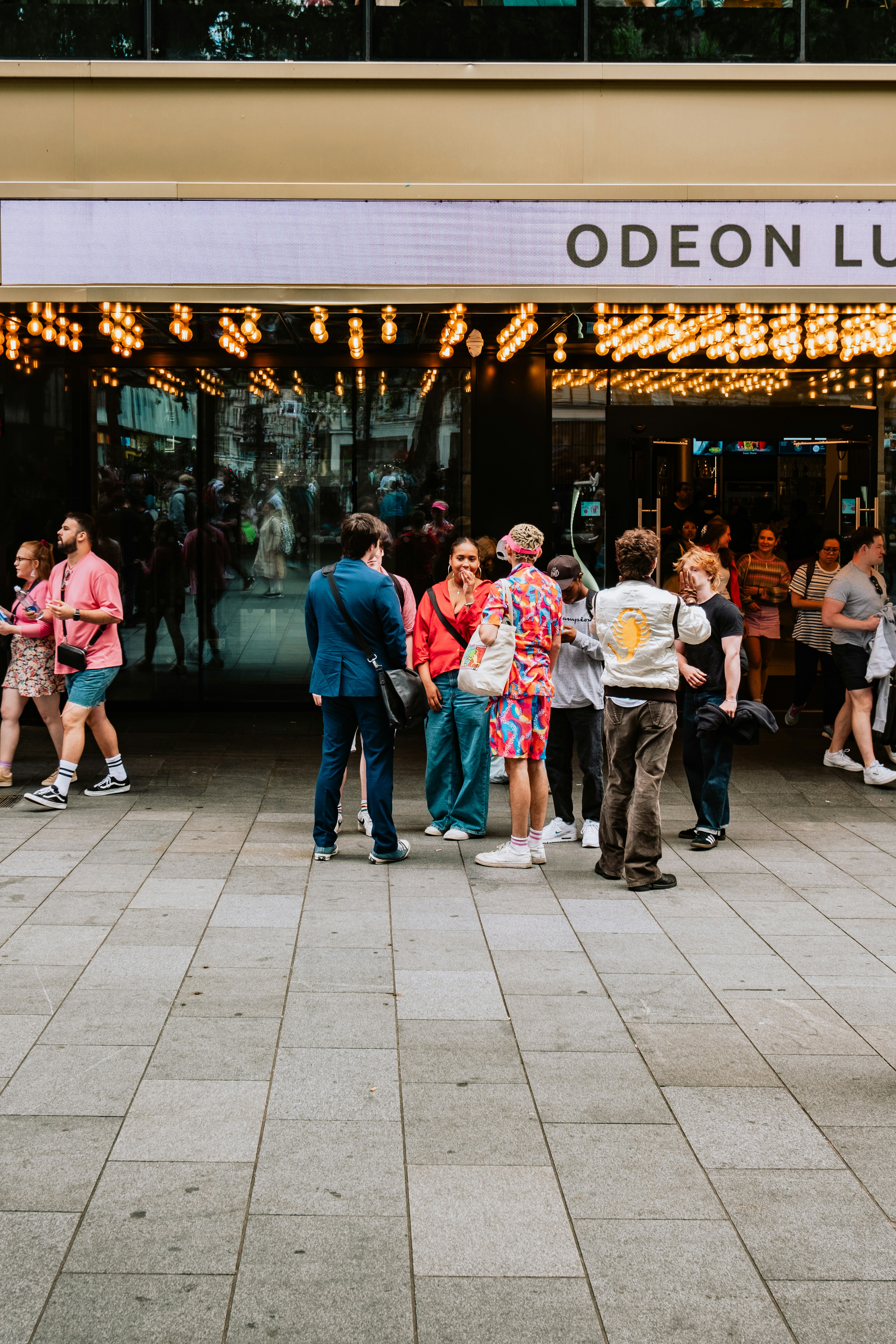 A group of people stand outside a cinema entrance adorned with numerous bright lights. The cinema's name 'Odeon Lux' is partially visible on the sign above the entrance. The individuals appear to be engaged in conversation, with a variety of colorful and casual outfits. The area is busy, with other people visible entering or exiting the venue. The scene suggests a lively, social atmosphere.