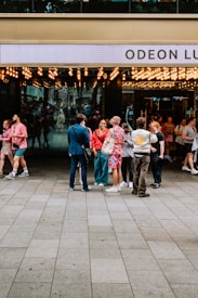 A group of people stand outside a cinema entrance adorned with numerous bright lights. The cinema's name 'Odeon Lux' is partially visible on the sign above the entrance. The individuals appear to be engaged in conversation, with a variety of colorful and casual outfits. The area is busy, with other people visible entering or exiting the venue. The scene suggests a lively, social atmosphere.